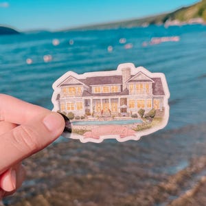 May include: A sticker featuring a watercolor illustration of a large house with a pool. The house has multiple windows and a dark roof. A pink banner at the bottom reads "Lakeside Beach". The sticker is held up against a blurred background of water.