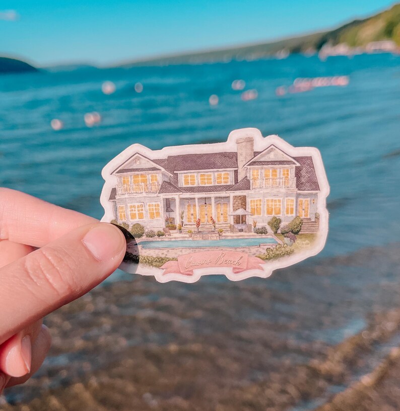 May include: A sticker featuring a watercolor illustration of a large house with a pool. The house has multiple windows and a dark roof. A pink banner at the bottom reads "Lakeside Beach". The sticker is held up against a blurred background of water.