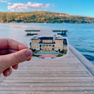 May include: A sticker featuring a detailed illustration of a large house with a pool, set against a backdrop of a lake and mountains. The house has multiple windows and a dark roof. A pink banner with cursive text is below the house.