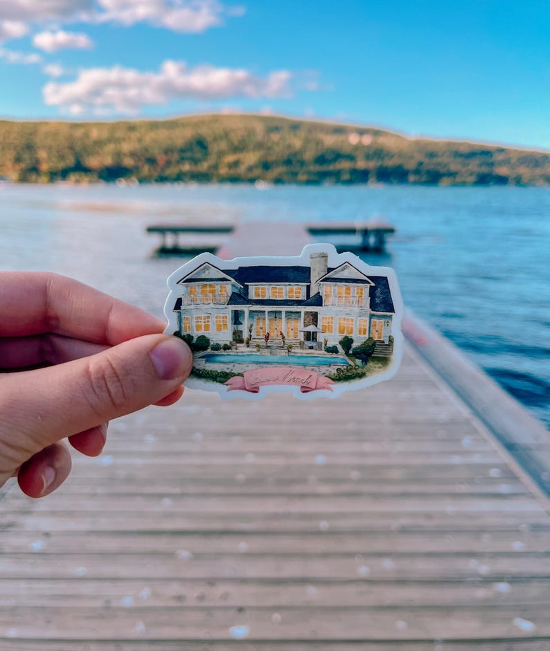 May include: A sticker featuring a detailed illustration of a large house with a pool, set against a backdrop of a lake and mountains. The house has multiple windows and a dark roof. A pink banner with cursive text is below the house.