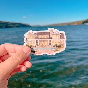 May include: A sticker featuring a watercolor illustration of a large white house with a dark roof, a swimming pool, and landscaping. The sticker is held against a backdrop of a blue lake and sky.