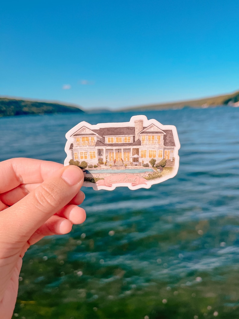 May include: A sticker featuring a watercolor illustration of a large white house with a dark roof, a swimming pool, and landscaping. The sticker is held against a backdrop of a blue lake and sky.