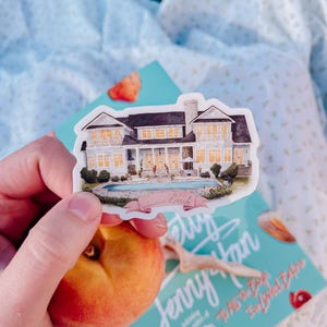 May include: A white sticker featuring a watercolor illustration of a large house with a pool. The sticker has a banner that reads "Sunset Beach". The sticker is held over a book and a peach.