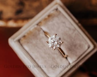 May include: A round, clear diamond engagement ring set in a gold band, displayed in a white velvet ring box. The box is sitting on a brown surface.  "GOLDEN PRO JEWELS GOLDEN PRO"