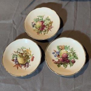 Puede incluir: Tres cuencos de postre de cerámica blanca con ribete dorado y diseño de frutas. Cada cuenco presenta una disposición de frutas diferente, que incluye uvas, peras, manzanas y bayas.