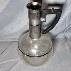 May include: A silver and glass pitcher with a black plastic handle. The pitcher has a round body and a tall, cylindrical neck.