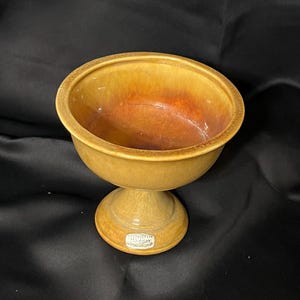 Op de afbeelding: Een gele keramische voetkom met een bruine glazuur. De kom heeft een ronde voet en een brede, ondiepe kom bovenop. De kom heeft een label met de tekst "Haeger Pottery, Made in USA".