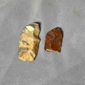 May include: Two flint arrowheads, one is a light brown color with white streaks, the other is a dark red color with white streaks.