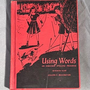 Puede incluir: Un libro rojo con una cubierta negra y el título "Using Words: An Enriched Spelling Program, Seventh Year" de Lillian E. Billington. La cubierta presenta una ilustración en blanco y negro de niños practicando tiro con arco.