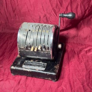 May include: Vintage black Paymaster Model V check writer with a silver-toned face and a black crank handle. The check writer has a series of numbered dials and a small lever.