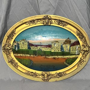 Puede incluir: Una imagen enmarcada de forma ovalada con un marco dorado y una imagen pintada de un palacio con un cielo azul y árboles verdes. El texto "Palace of Versailles" está impreso en la parte inferior de la imagen.