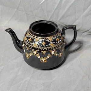 以下が含まれることがあります： 白、オレンジ、金色の花柄が施された黒いティーポット。ティーポットは丸いボディと湾曲した注ぎ口を持っています。