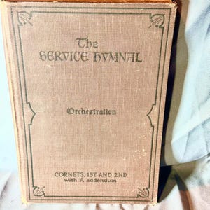 May include: The cover of a vintage book titled "The Service Hymnal: Orchestration, Cornets 1st and 2nd with A addendum". The book is brown with a green border and a decorative design on the front cover.