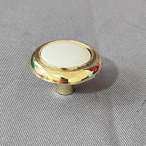 May include: A round, white ceramic knob with a gold metal base. The knob has a raised, circular design around the edge.