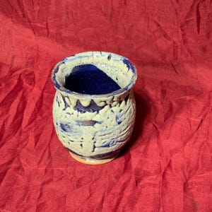 May include: A small, white ceramic vase with a blue glaze and a textured surface. The vase has a rounded body and a flared rim.