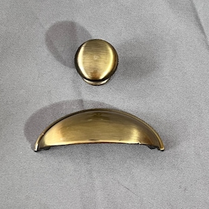 May include: Two antique brass cabinet hardware pieces. One is a round knob, and the other is a half-moon pull. The hardware has a warm, aged finish, suggesting a vintage or classic style. The pieces are set against a neutral grey background.