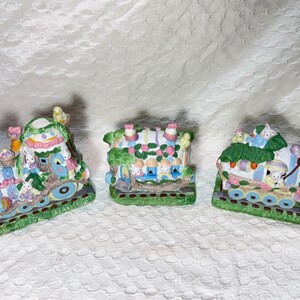 May include: Three ceramic Easter bunny train cars decorated with pastel colors, flowers, and green leaves. Each car features a bunny with a different expression.