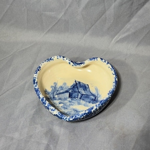 May include: A heart-shaped ceramic dish with a blue and white glaze. The dish features a hand-painted scene of a small house in a wooded area.