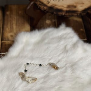 May include: A white agate pendulum with a silver chain and black beads. The pendulum is hanging on a white fluffy surface.