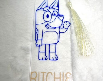 May include: A clear acrylic bookmark with a blue outline of a cartoon dog. The dog is wearing a blue shirt and has a white tassel. The bookmark has the name "RITCHIE" printed in gold letters.