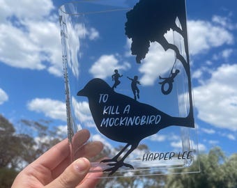 Puede incluir: Funda de libro de acrílico transparente con una silueta negra de un pájaro y niños jugando bajo un árbol. El texto "TO KILL A MOCKINGBIRD" y "HARPER LEE" están impresos en la funda. El fondo es un cielo azul con nubes.
