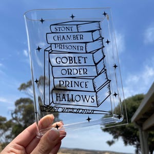 以下が含まれることがあります： 透明なアクリル製のトレイに、黒い輪郭で「ストーン」、「チェンバー」、「プリズナー」、「ゴブレット」、「オーダー」、「プリンス」、「ハロウズ」というタイトルの書物が積み重ねられています。本の真下には、黒い輪郭の杖が描かれています。