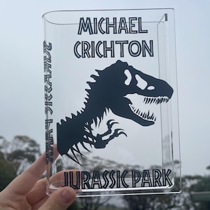 Może przedstawiać: Przezroczysty, akrylowy stojak w kształcie książki z napisem "MICHAEL CRICHTON" i "JURASSIC PARK" oraz czarną sylwetką dinozaura. Grzbiet książki zawiera napis "JURASSIC PARK" w pionie. Stojak jest trzymany na tle pochmurnego nieba.