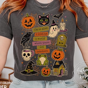 May include: A charcoal gray t-shirt featuring a colorful Halloween-themed graphic print. The design includes a pumpkin, black cat, ghost, raven, skull mug, frog, witch hat, tombstone, and house with the text, I'm so glad I live in a world where there are octobers.