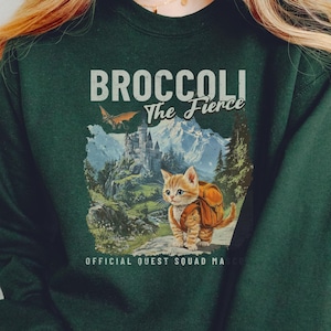 K&ouml;nnte beinhalten: Ein dunkelgr&uuml;nes Sweatshirt mit einem Aufdruck eines Katers mit Rucksack und dem Text "Broccoli The Fierce" und "Official Quest Squad Member".