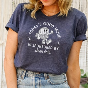 Puede incluir: Una camiseta azul marino con un gráfico blanco de una computadora retro sosteniendo un teclado y dando un pulgar hacia arriba. El texto en la camiseta dice "Today's Good Mood Is Sponsored By Clean Data."