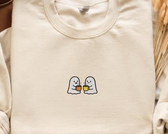 May include: Sand-coloured sweatshirt with a crew neck and long sleeves. The front has an embroidered design of two ghost figures holding coffee mugs. The word "SAND" is printed at the bottom left.