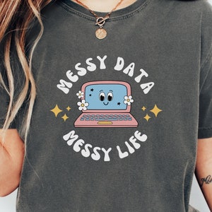 Könnte beinhalten: Ein dunkelgraues T-Shirt mit einer Retro-Grafik eines Laptops mit einem lächelnden Gesicht. Der Text "Messy Data Messy Life" ist in einer wellenförmigen Schrift um den Laptop herum geschrieben.