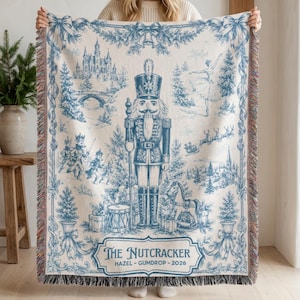 May include: A woven throw blanket featuring a blue and cream toile-style print of a nutcracker, a castle, a ballerina, and holiday motifs. The text reads THE NUTCRACKER HAZEL - GUMDROP - 2026. The blanket has a fringed edge and a decorative border.