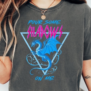 May include: Dark grey t-shirt with a blue and pink dragon graphic. The design includes the text "Pour Some Shadows On Me" inside a triangle. The shirt is made of a soft, comfortable material.