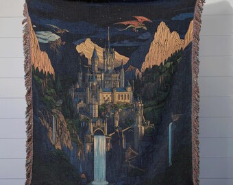 May include: A woven tapestry throw blanket featuring a dark blue background with a detailed illustration of a castle nestled in a mountain range with a waterfall cascading down the side. The castle is illuminated with warm yellow lights and there are two red dragons flying in the sky above.
