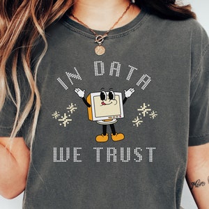 Può includere: Una maglietta grigio scuro con un design a pixel che dice "In Data We Trust". Il design presenta un monitor di computer di cartone animato con un viso sorridente e le braccia allargate. Il monitor del computer è giallo e ha uno schermo bianco. Ci sono piccole stelle bianche intorno al monitor del computer.