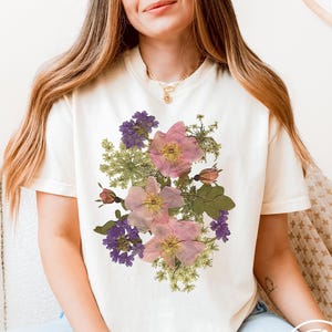 Woman wearing an ivory Comfort Colors T-shirt with a vintage-style floral design featuring pressed-style botanical illustrations of pink wild roses, deep purple larkspur flowers, and delicate green Queen Anne’s lace foliage. The soft, natural color palette and hand-drawn appearance of the design give the shirt a romantic, cottagecore feel. She is sitting casually in a light, cozy setting with layered gold necklaces and soft waves in her hair. Lilio Creations branding in lower right.