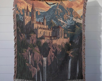 May include: A woven tapestry throw blanket featuring a fantasy castle nestled in a mountain range with waterfalls cascading down the cliffs. The castle has a pointed roof and multiple towers. The sky is a vibrant orange and pink sunset.