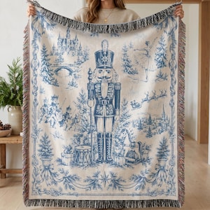 May include: A woven throw blanket featuring a blue and cream toile-style pattern of a central nutcracker figure surrounded by a castle, a ballerina, a rocking horse, and Christmas trees, finished with a decorative fringed edge on all sides.