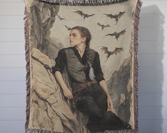 May include: A woven tapestry throw blanket features a woman with long brown hair and a dark brown leather vest. She is sitting on a rock with a background of a rocky landscape and several bats flying in the air.