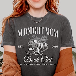 以下が含まれることがあります： 白い文字で「Midnight Mom Book Club」と書かれた濃いグレーのTシャツ。デザインには、本を積み重ねたもの、コーヒーカップ、眼鏡が描かれている。「Est. 2016」と「Reading Past Bedtime Since Forever」の文字も含まれている。
