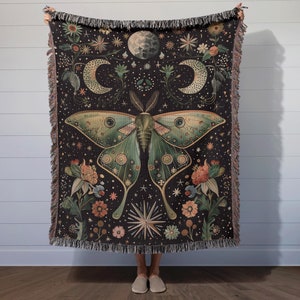 May include: A woven tapestry throw blanket with a dark background featuring a green and gold moth with intricate details, surrounded by flowers and stars. The moth's wings are spread wide, and the design is reminiscent of a celestial night sky.