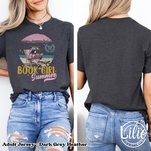 May include: Dark grey heather adult jersey t-shirt with a graphic of a raccoon wearing sunglasses and reading a book on a beach chair under an umbrella. The text "Book Girl Summer" is printed on the shirt.