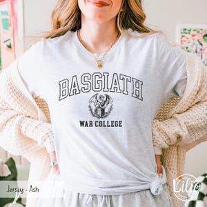 K&ouml;nnte beinhalten: Ein hellgraues T-Shirt mit dem Text "BASGIATH WAR COLLEGE" in Schwarz und einem Drachenemblem. Das Shirt besteht aus einem weichen Jersey-Material.