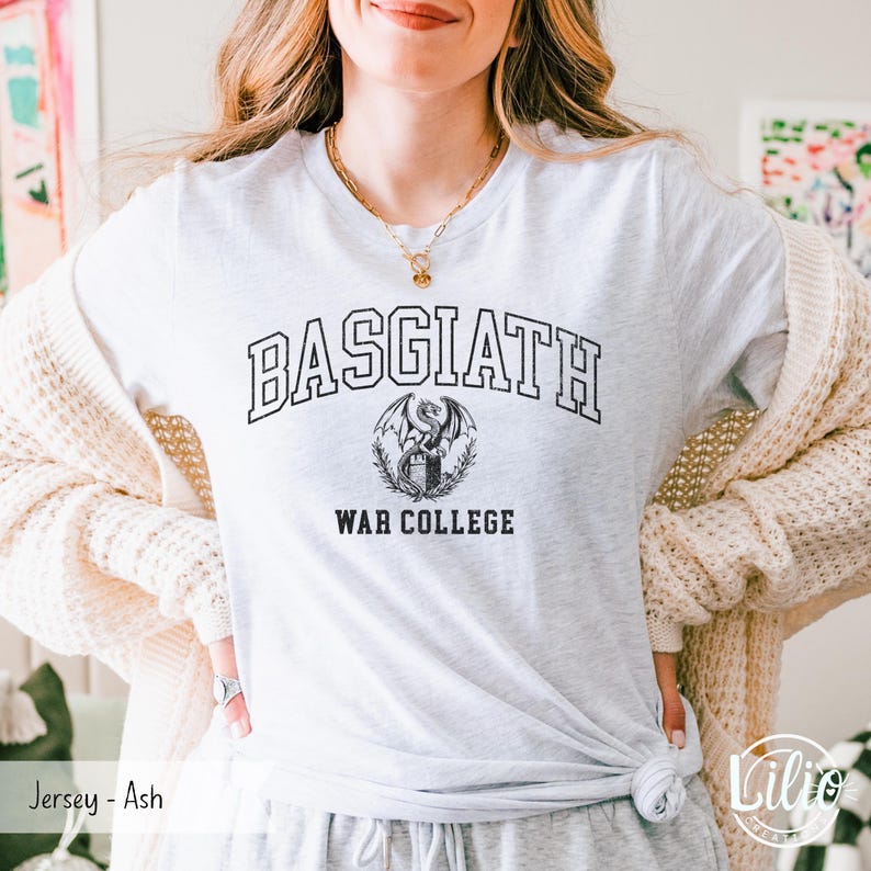 K&ouml;nnte beinhalten: Ein hellgraues T-Shirt mit dem Text "BASGIATH WAR COLLEGE" in Schwarz und einem Drachenemblem. Das Shirt besteht aus einem weichen Jersey-Material.