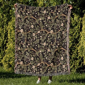 Pu&ograve; includere: Una coperta tessuta con sfondo scuro e motivo floreale con fiori bianchi, foglie verdi e rami marroni. La coperta ha un bordo sfrangiato ed &egrave; tenuta su uno sfondo verde.