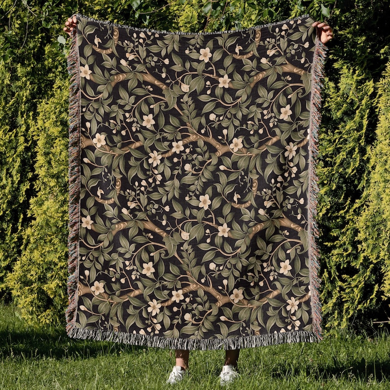 Pu&ograve; includere: Una coperta tessuta con sfondo scuro e motivo floreale con fiori bianchi, foglie verdi e rami marroni. La coperta ha un bordo sfrangiato ed &egrave; tenuta su uno sfondo verde.