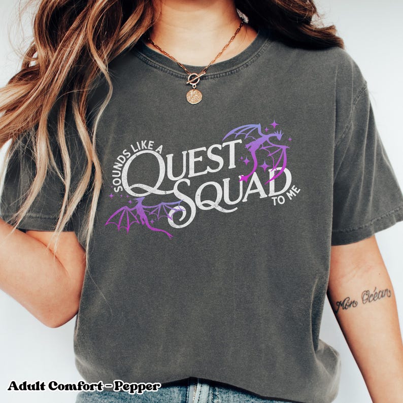 以下が含まれることがあります： グレーのTシャツに、紫と白のグラフィックがプリントされています。「Sounds like a Quest Squad to me」と書かれており、2匹のドラゴンと星が描かれています。