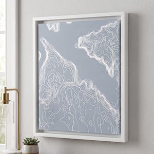 May include: A square framed wall art piece featuring a light gray background with white topographic contour lines. The artwork is encased in a simple white frame, displaying abstract elevation patterns that resemble a map of a coastline.