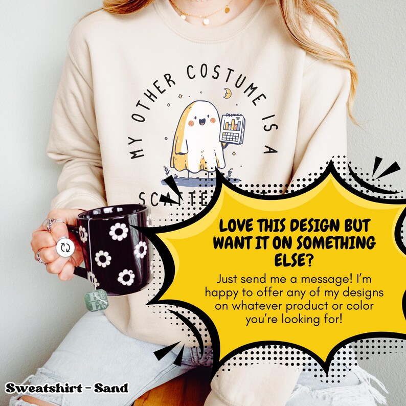 Pode incluir: Camisola cor de areia com um gr&aacute;fico de fantasma segurando um gr&aacute;fico e o texto "My Other Costume Is A Scante". Uma caneca preta com desenhos de flores brancas &eacute; segurada em primeiro plano. Uma bolha amarela diz "LOVE THIS DESIGN BUT WANT IT ON SOMETHING ELSE?"